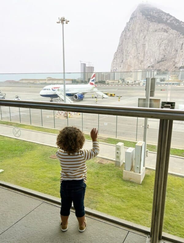 A young child in a striped shirt looking out an airport window at a British Airways airplane with a large mountain Gibraltar in the background.