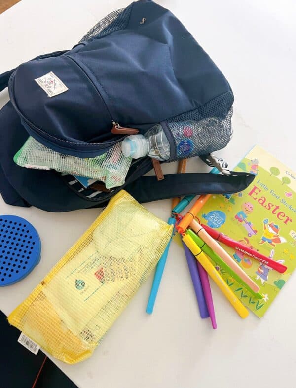 A dark blue Joules backpack on a white counter surrounded by travel activities, including Stabilo Cappi pens, a small water bottle, and an Usborne sticker book.