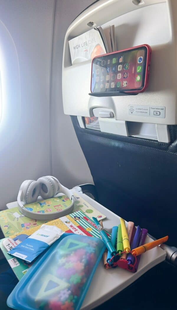 An airplane tray table set up with wireless headphones, a sticker book, Stabilo pens, disposable toilet seat covers, and a smartphone mounted on a seatback tablet stand.