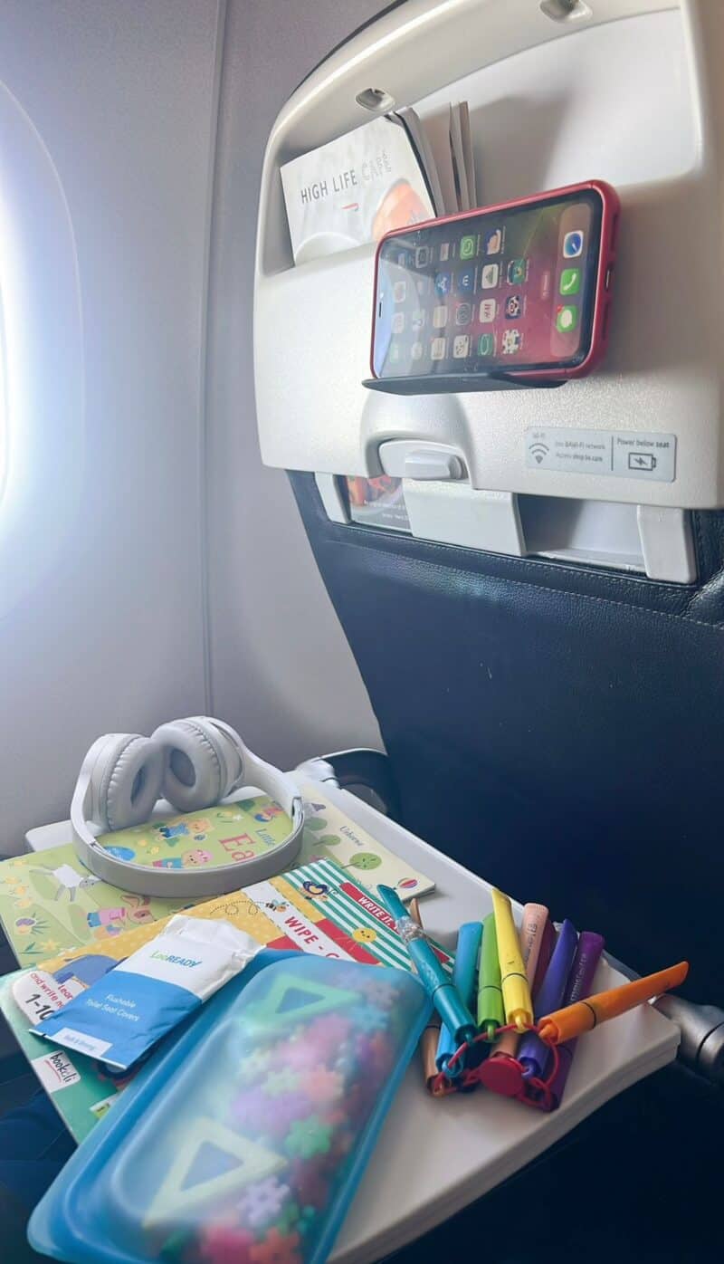 An airplane tray table set up with wireless headphones, a sticker book, Stabilo pens, disposable toilet seat covers, and a smartphone mounted on a seatback tablet stand.