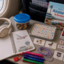 Plane activities for 5–7 year olds including Uno, letter games, notebook, Stabilo Cappi pens, Cat's Cradle string, headphones and snacks on an airplane tray table.